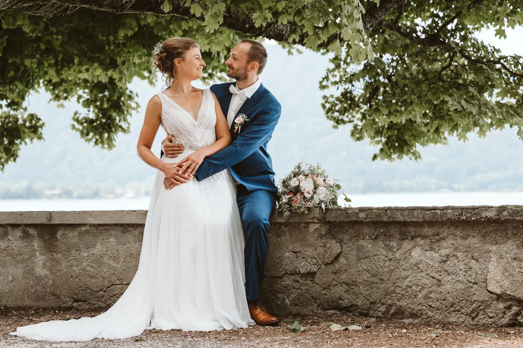 Romantische Hochzeitsfotos am Tegernsee. Brautpaar sitzt auf einer Steinmauer und blickt sich verliebt in die Augen, eine authentische Momentaufnahme aus der Hochzeitsreportage, fotografiert von Ihrem Hochzeitsfotograf Olching.