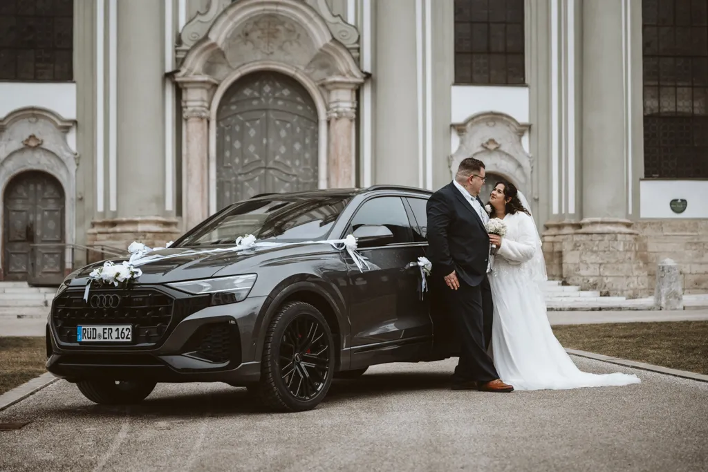 Stilvoller Moment am Hochzeitswagen Modernes Brautpaar-Shooting am Kloster Fürstenfeld: Braut und Bräutigam lehnen entspannt an ihrem Hochzeitsauto und sehen sich an.