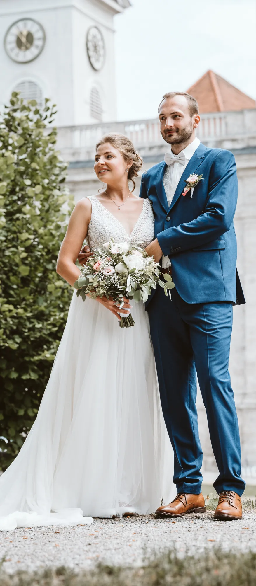 Der Bräutigam im blauen Anzug umarmt seine Braut im weißen Kleid von hinten, sie stehen vor der Kirche am Tegernsee. Eine emotionale Aufnahme, die ich als Ihr Fotograf für authentische Hochzeiten festgehalten habe.