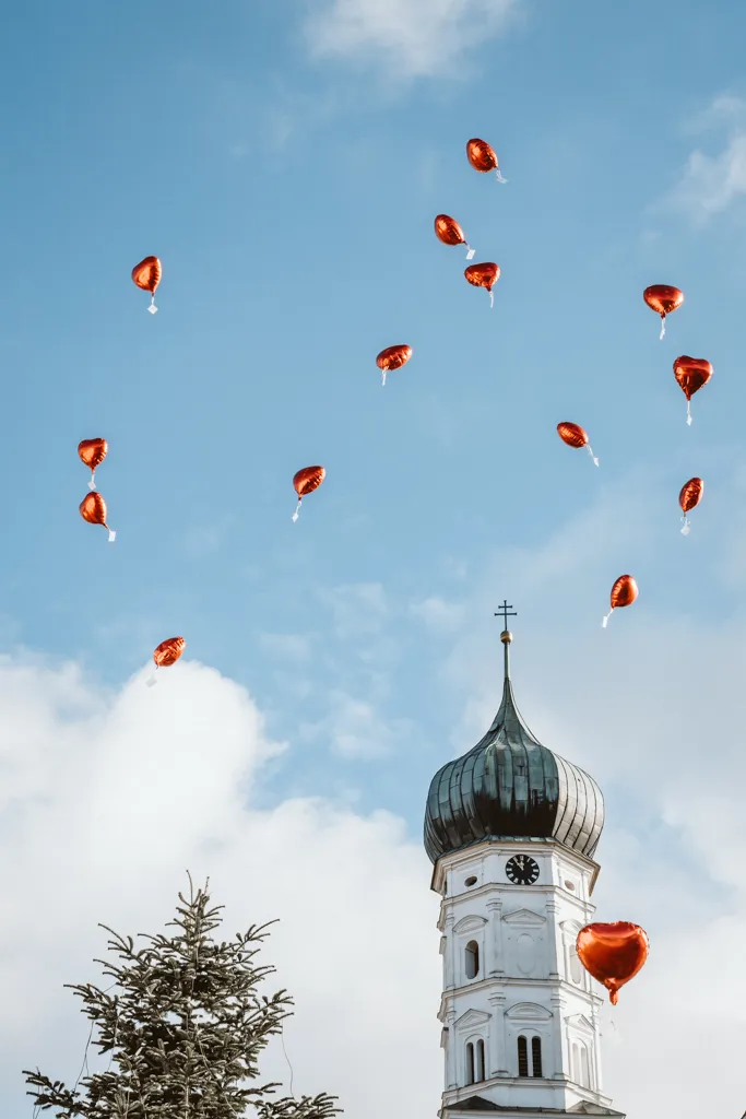 Auf dem Weg in den Himmel: Rote Herzluftballons vor malerischer Kirche. Rote Herzluftballons steigen vor einer Kirchturmspitze in den Himmel.