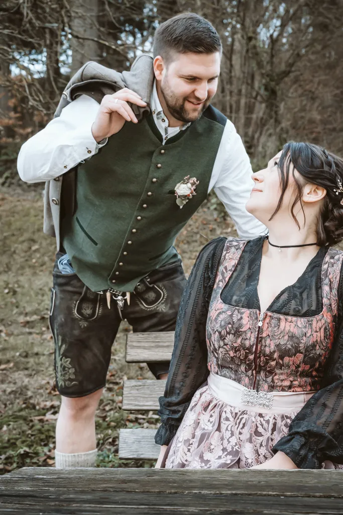 Lässige Eleganz: Brautpaar beim Fotoshooting. Bräutigam steht neben sitzender Braut, Jacke lässig über der Schulter, Blick auf sie gerichtet.