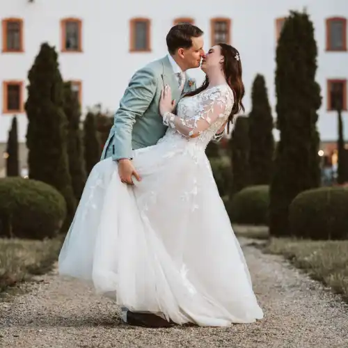 Ich habe das Hochzeitspaar im blauen Oldtimer fotografiert, wobei die Braut den Brautstrauß zum Fenster hinaus hält und winkt. Eine stimmungsvolle Aufnahme von mir als Hochzeitsfotograf Olching, die die freudige Fahrt dokumentiert.